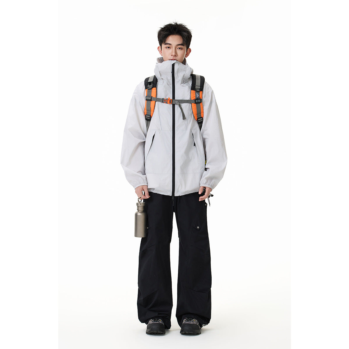 Model wearing a light gray high-collar zip jacket with a backpack, black wide-leg trousers, and holding a metal bottle.