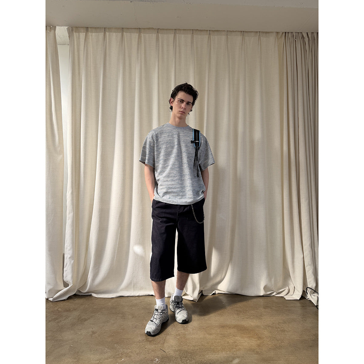 Model wearing a gray horizontally striped T-shirt, black knee-length shorts and a shoulder bag, facing forward.