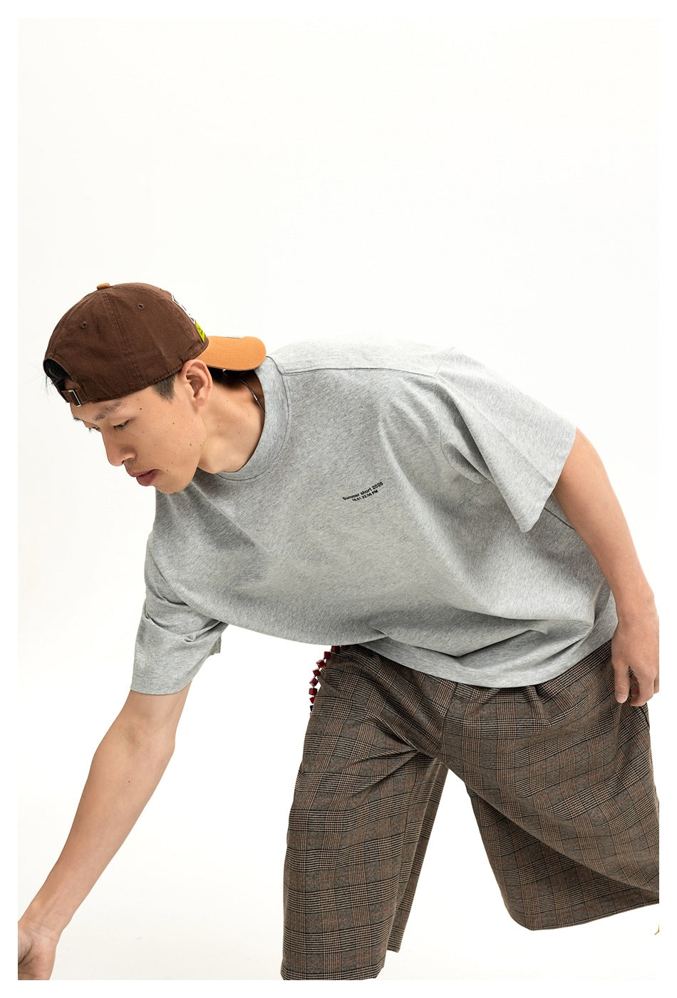 A model leans forward in a light gray oversized T-shirt and brown plaid shorts, showing the small chest print.