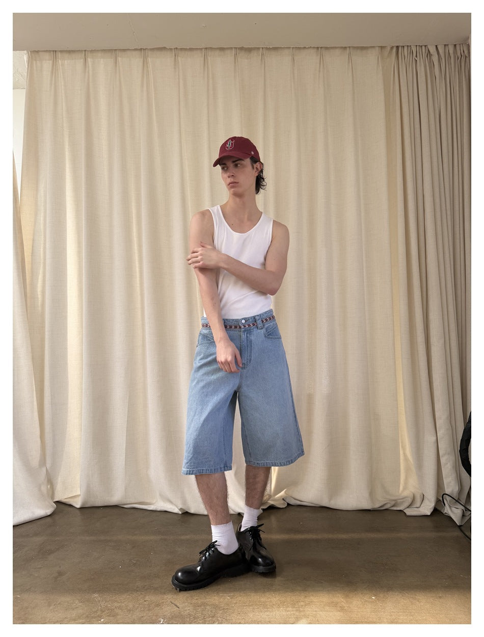 Model in light blue denim knee-length shorts, white tank top, red cap and black shoes, posed against a cream curtain