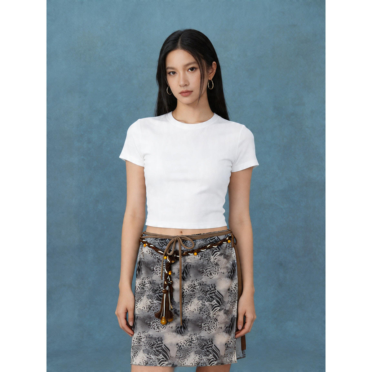 Model wearing a white cropped T-shirt and gray animal-print mini skirt with brown braided trim and a tie waist.