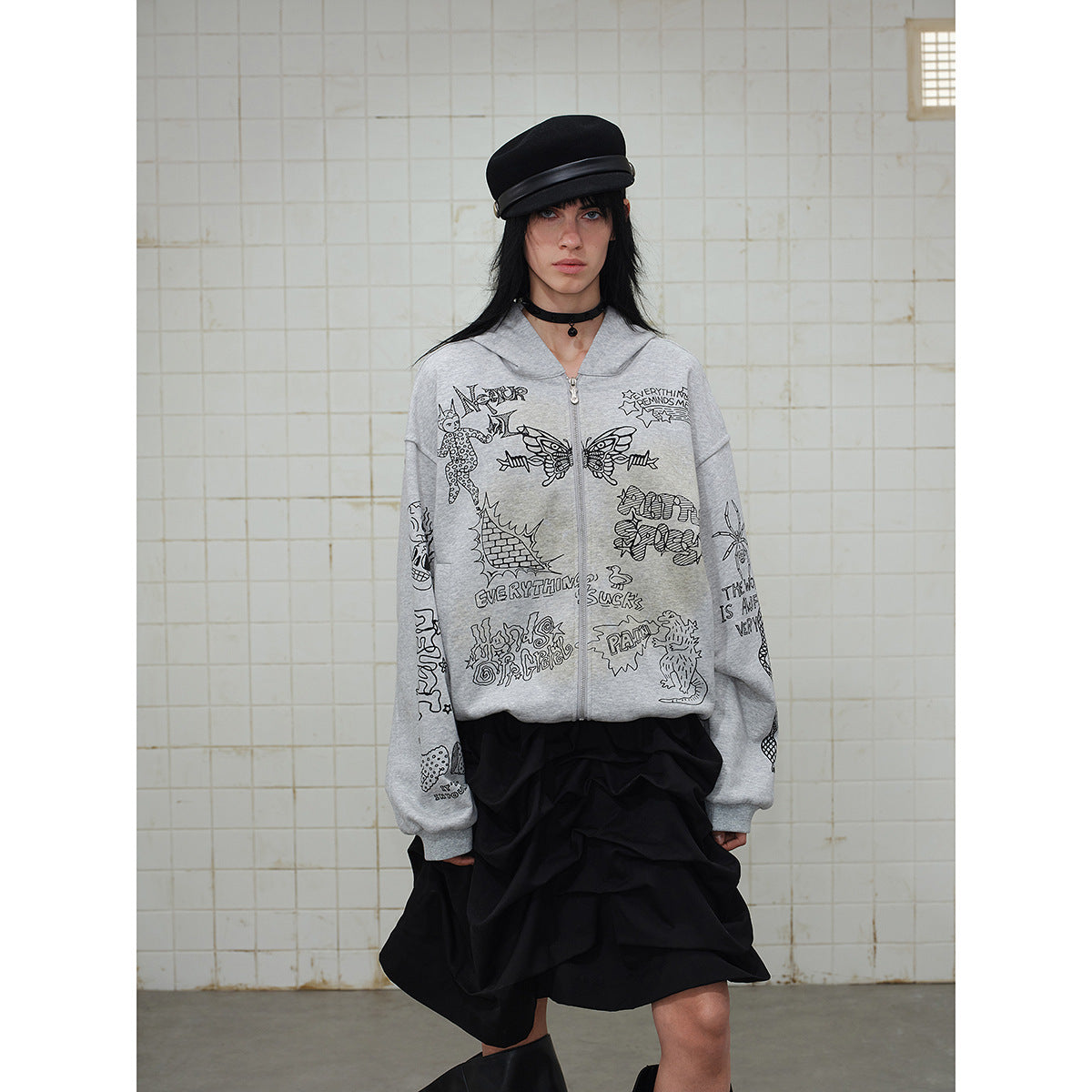 Front view of a model in a gray zip-up hoodie covered with black doodle graphics and a black ruffled skirt.