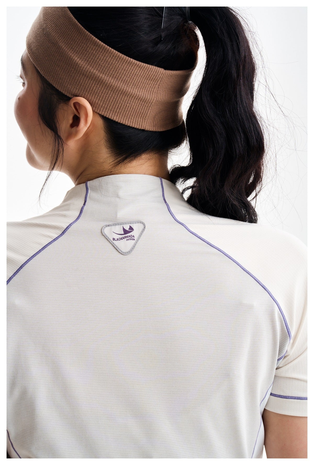 Rear view of a model wearing an ivory athletic shirt with lavender piping and a taupe headband.