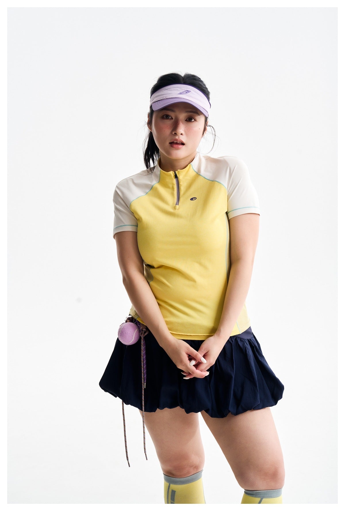 Model wearing a yellow and ivory zip-neck athletic top with a navy bubble skirt and purple visor.