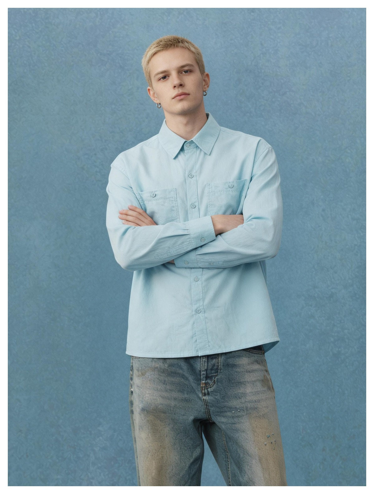 Model with arms crossed wearing a light blue button-up shirt with chest pockets and faded jeans against a blue backdrop
