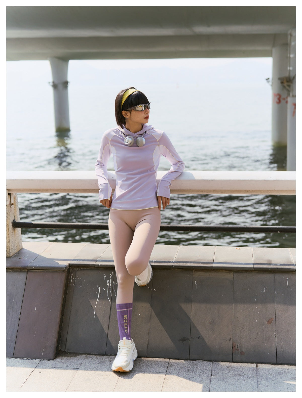Woman wearing the light purple hoodie with beige leggings, leaning on a waterfront railing