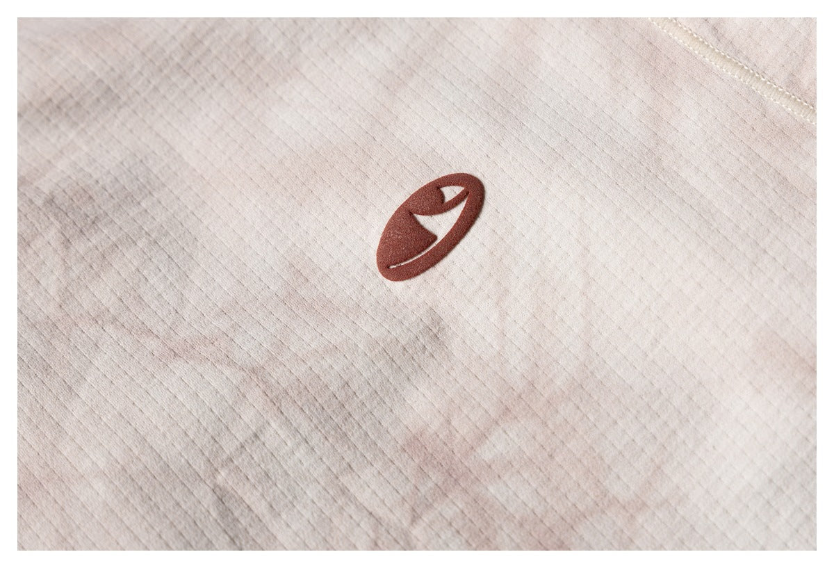 Close-up of a small dark brown emblem on the beige shirt’s mottled fabric.