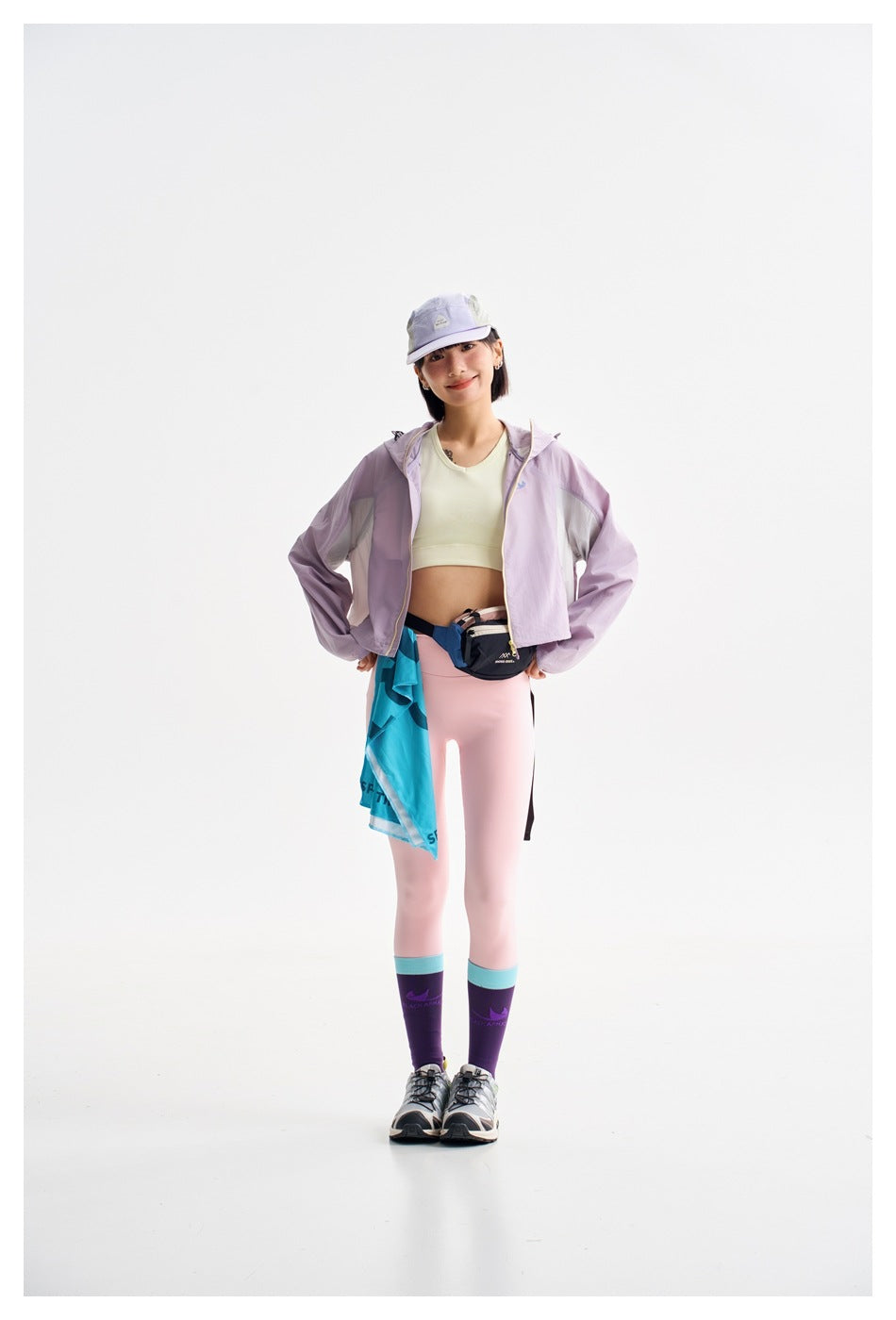 Model in a lilac cropped hooded jacket, pale crop top, pink leggings, and a lavender cap, shown from the front.