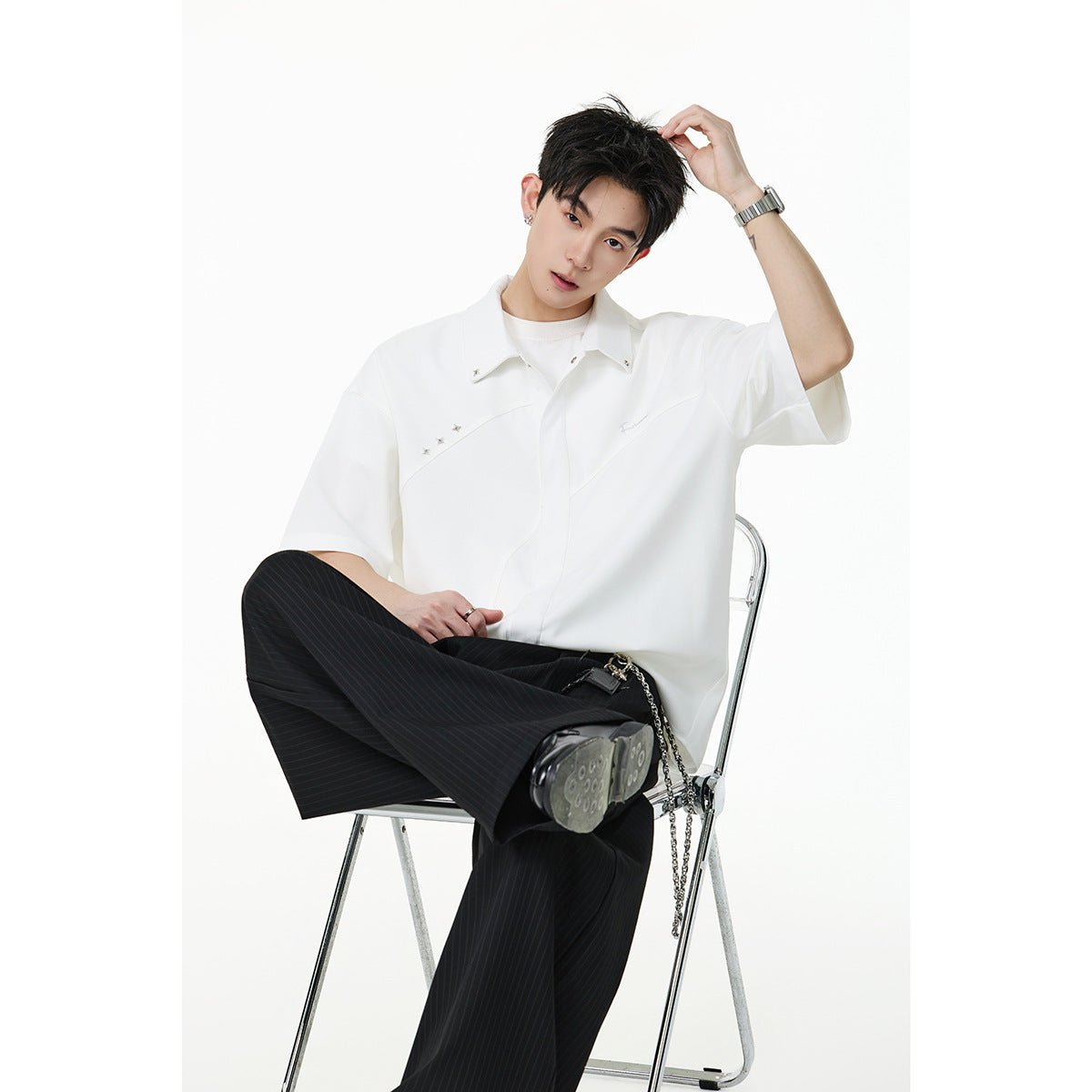 Seated model wearing a white short-sleeve button-up shirt with silver accents and black pinstripe trousers.