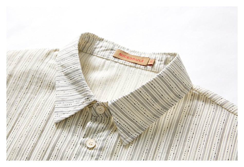 Close-up of an ivory shirt’s pointed collar and button placket, patterned with fine black dotted stripes.