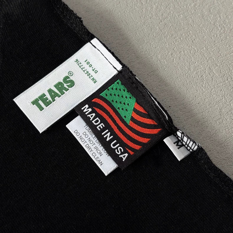 Close-up of tags on the Denim Tears Black Cotton Wreath All-Over Print T-Shirt. One tag shows Denim Tears?; another has a green and red American flag with MADE IN USA above care instructions on this oversized graphic tee.