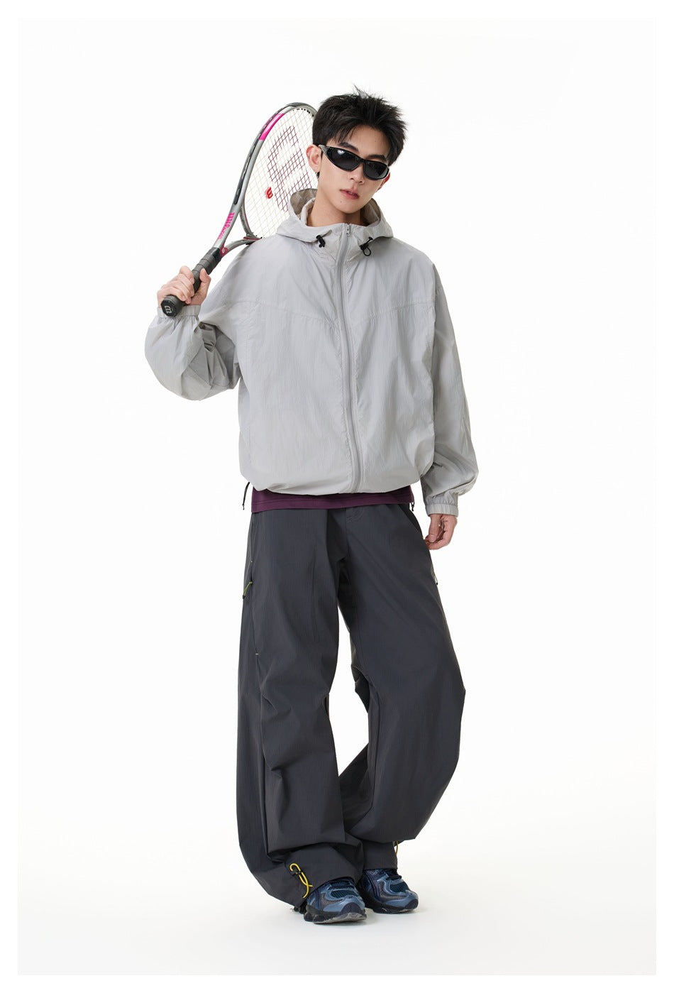 Full-length model wearing a light gray hooded zip jacket and dark trousers, holding a tennis racket.