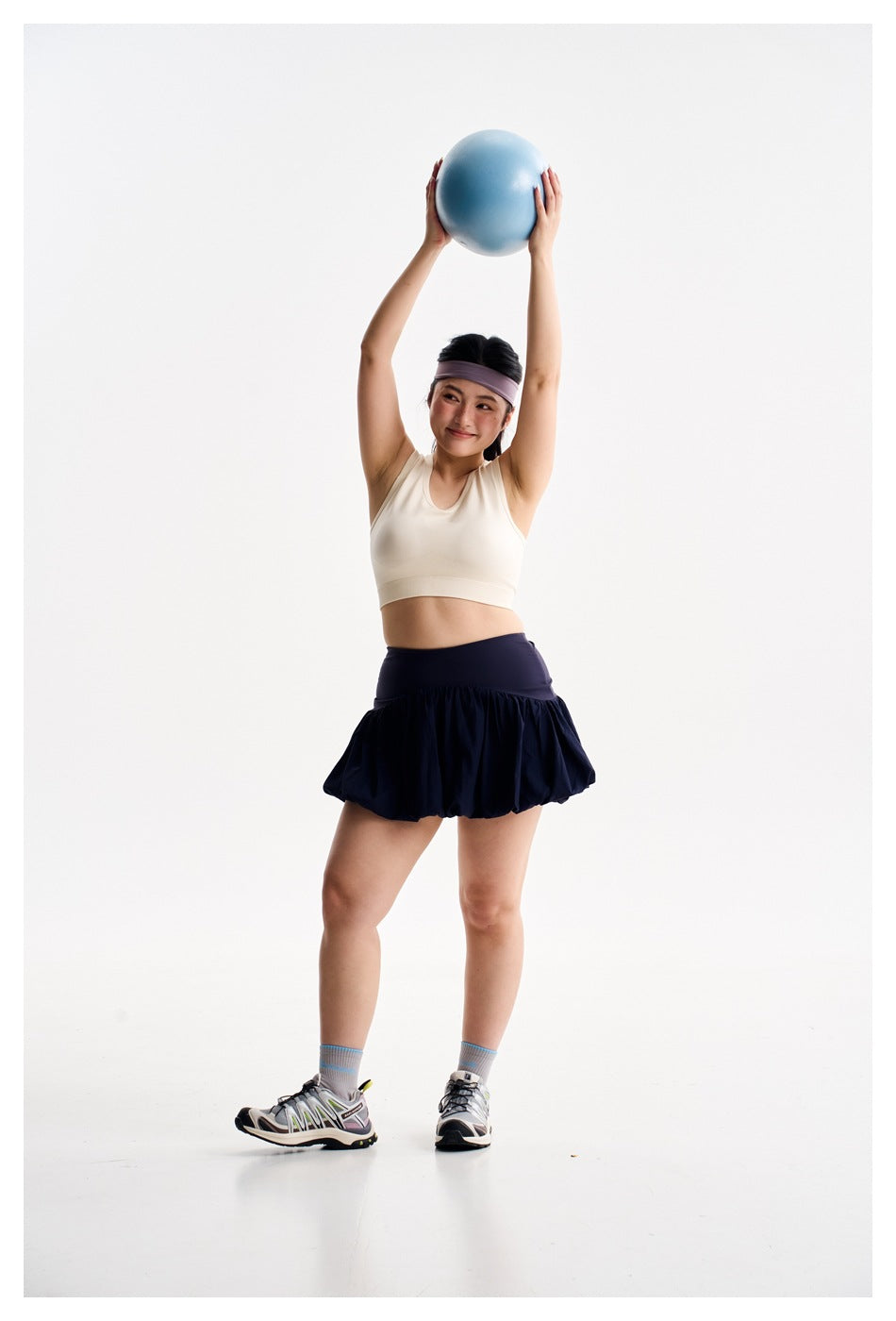 Woman in an ivory crop top and navy bubble-hem athletic skirt, lifting a blue exercise ball overhead.