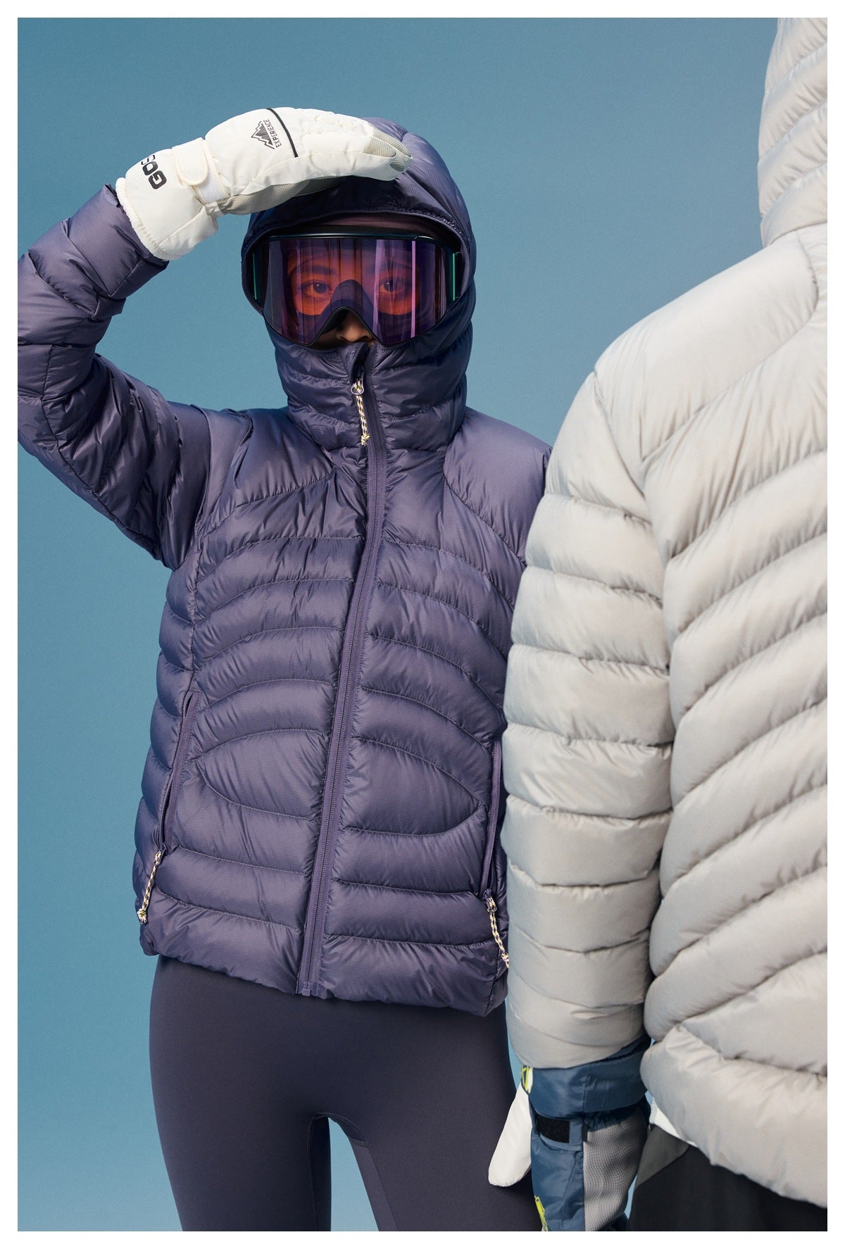 Person wearing a zipped lavender quilted hooded puffer jacket and ski goggles; another person partially blocks the right