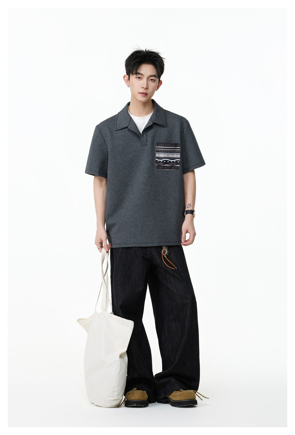 Model wearing a charcoal short-sleeve polo with a patterned chest pocket, paired with wide black jeans.