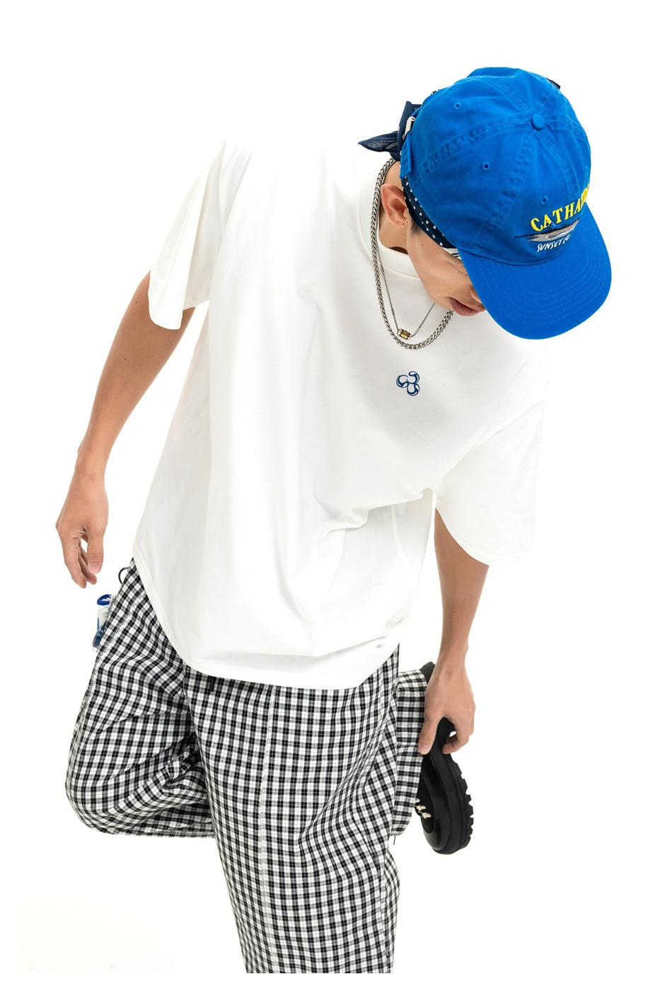 Model in an oversized white T-shirt with a small chest emblem, black-and-white check trousers, and a bright blue cap.