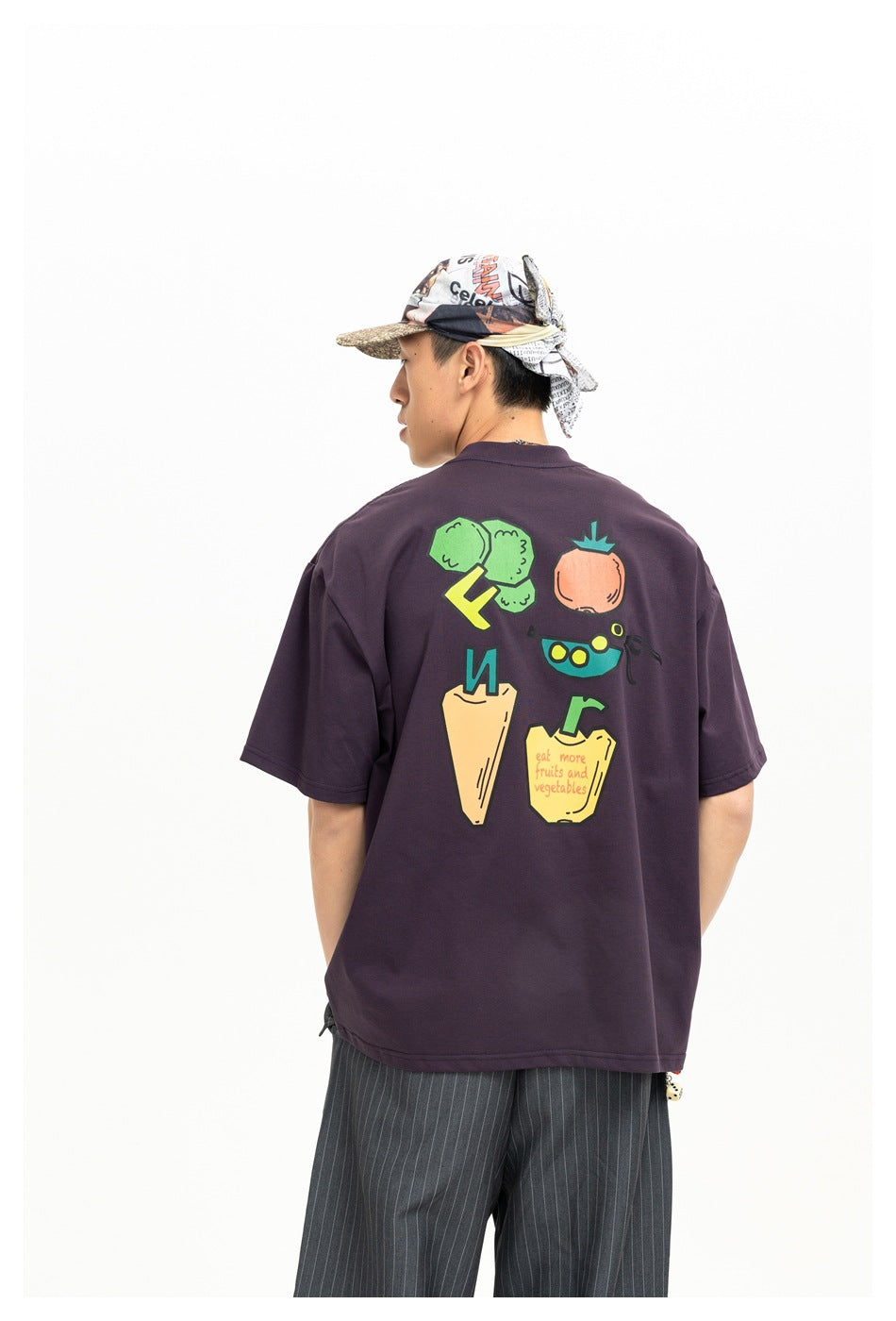 Back view of a model wearing a dark plum T-shirt with colorful fruit and vegetable graphics and English text.