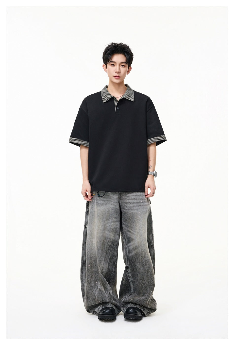 Front view of a model wearing a loose black polo with checked collar and sleeve cuffs, paired with wide gray jeans.