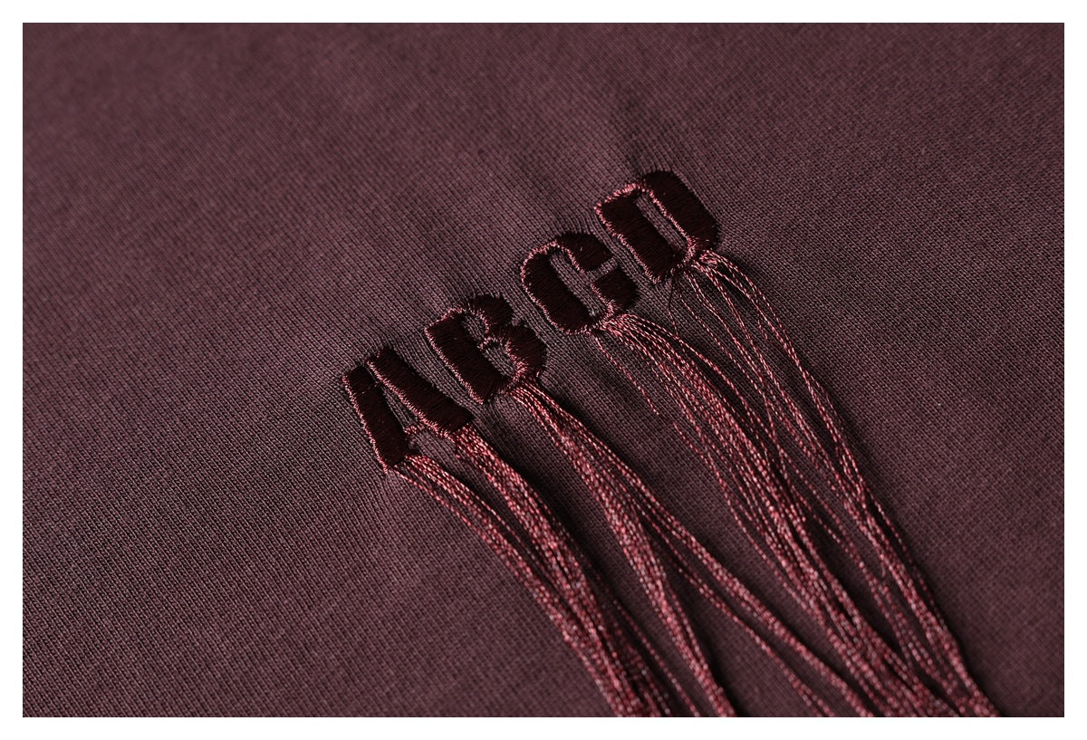 Close-up of dark burgundy fabric with raised dark embroidery and long burgundy threads trailing from the lettering.