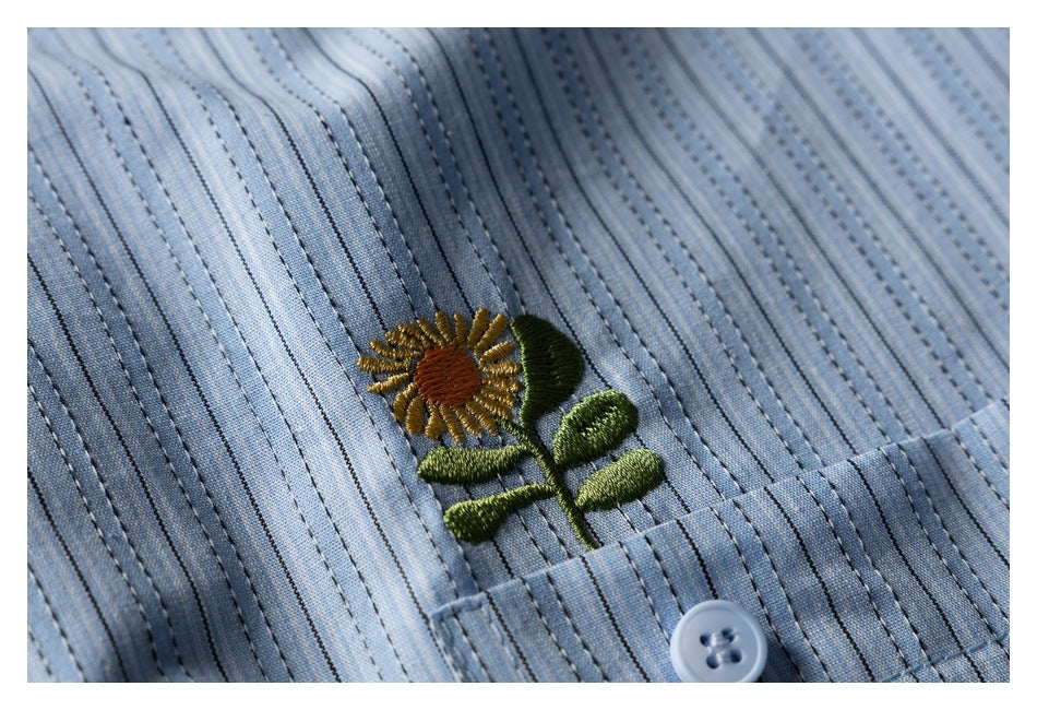 Close-up of a small embroidered sunflower and button on a light blue striped shirt.