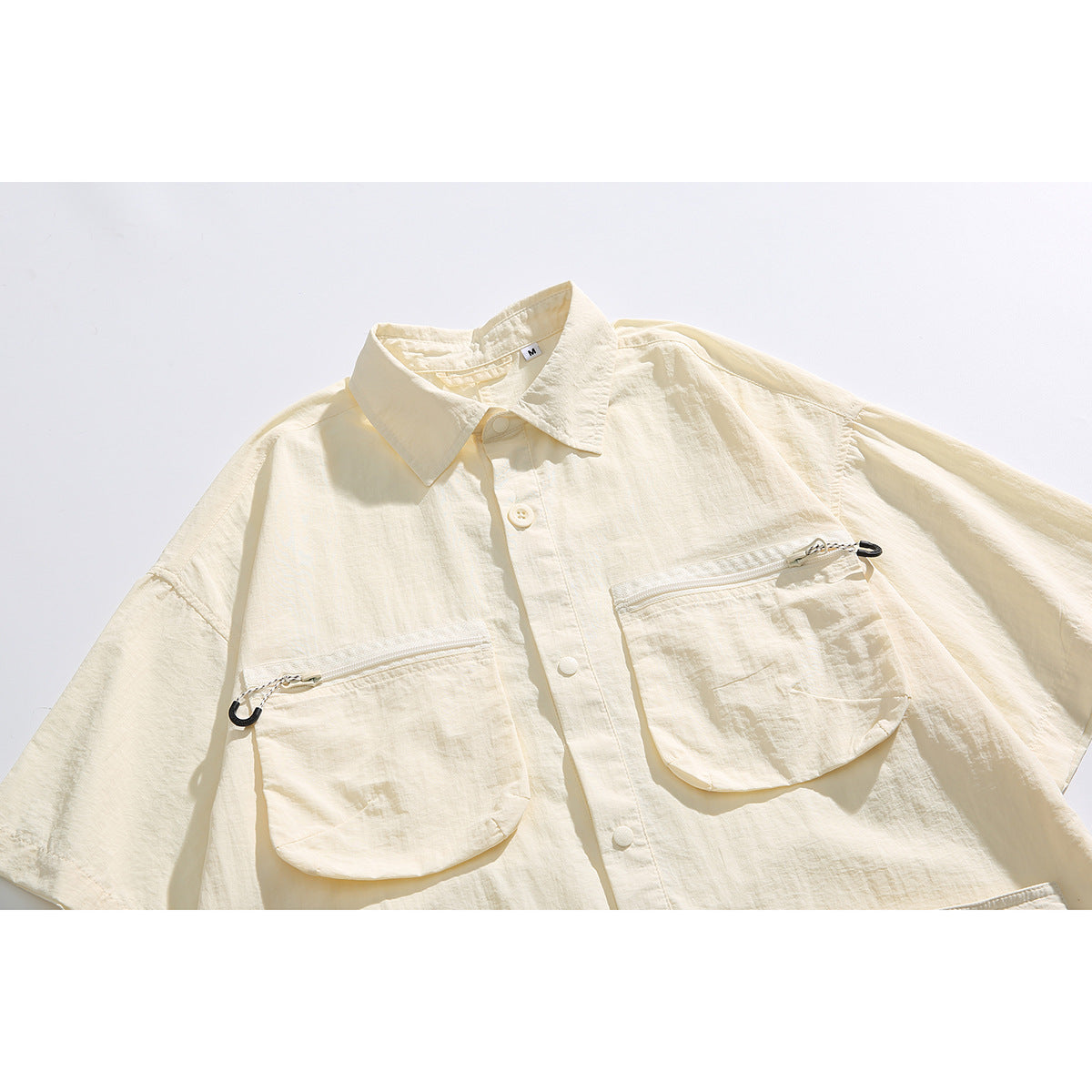 Close front view of an ivory short-sleeve collared shirt with buttons and two large zippered chest pockets.