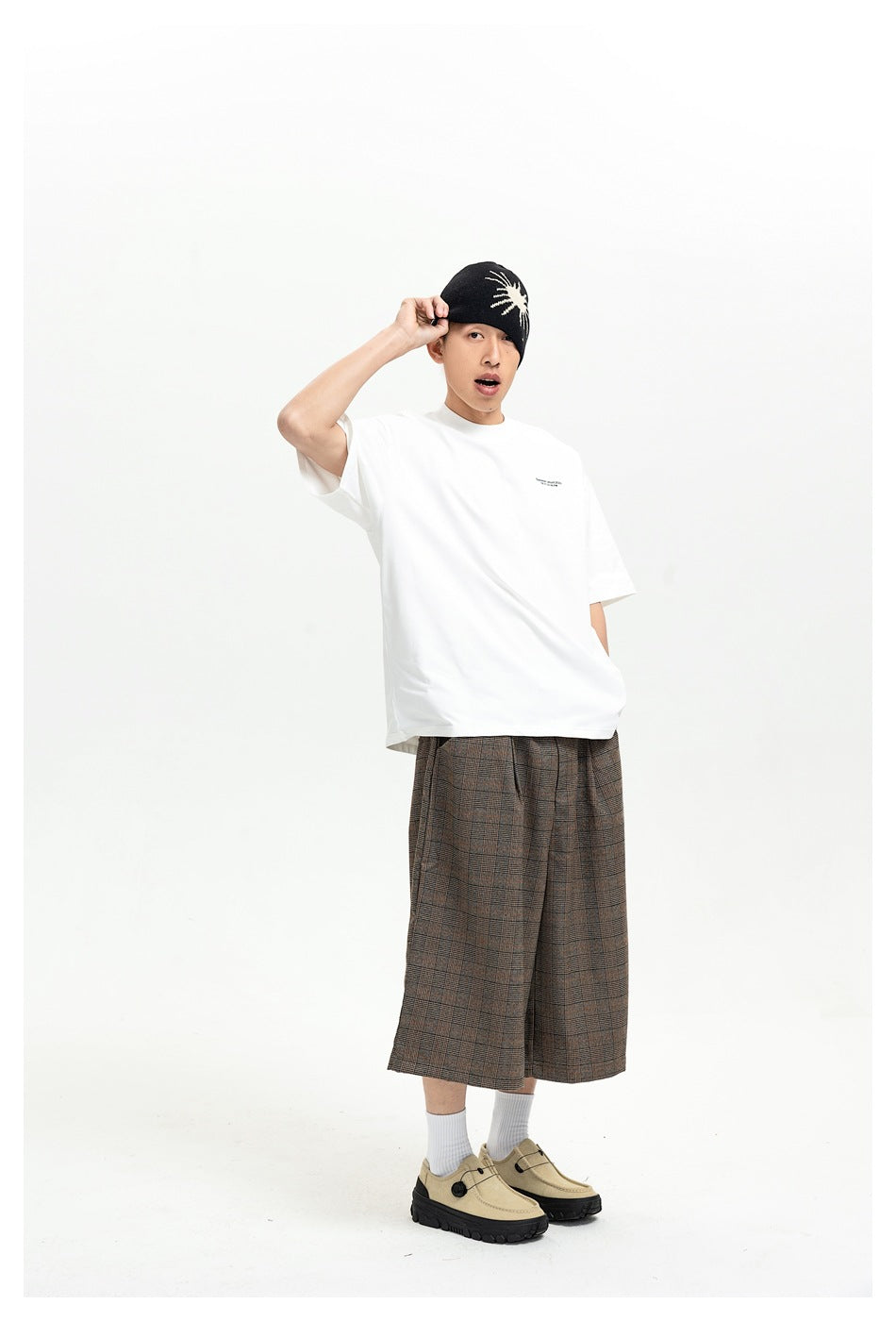 Model wearing a white oversized T-shirt with plaid wide-leg shorts, a black beanie and beige shoes, full-length view