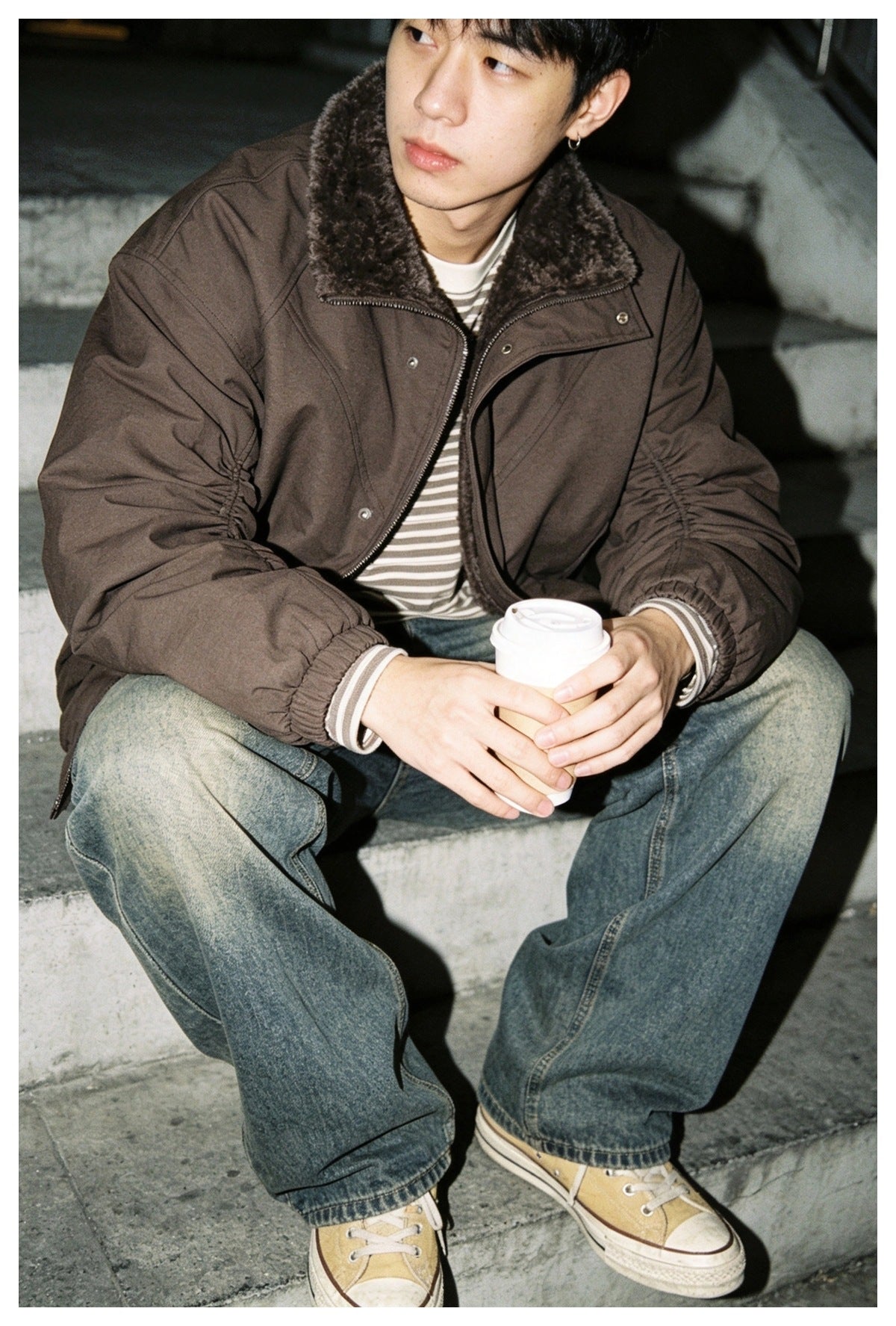 Man seated on steps wearing an open brown jacket with pile collar over a striped top, jeans and sneakers