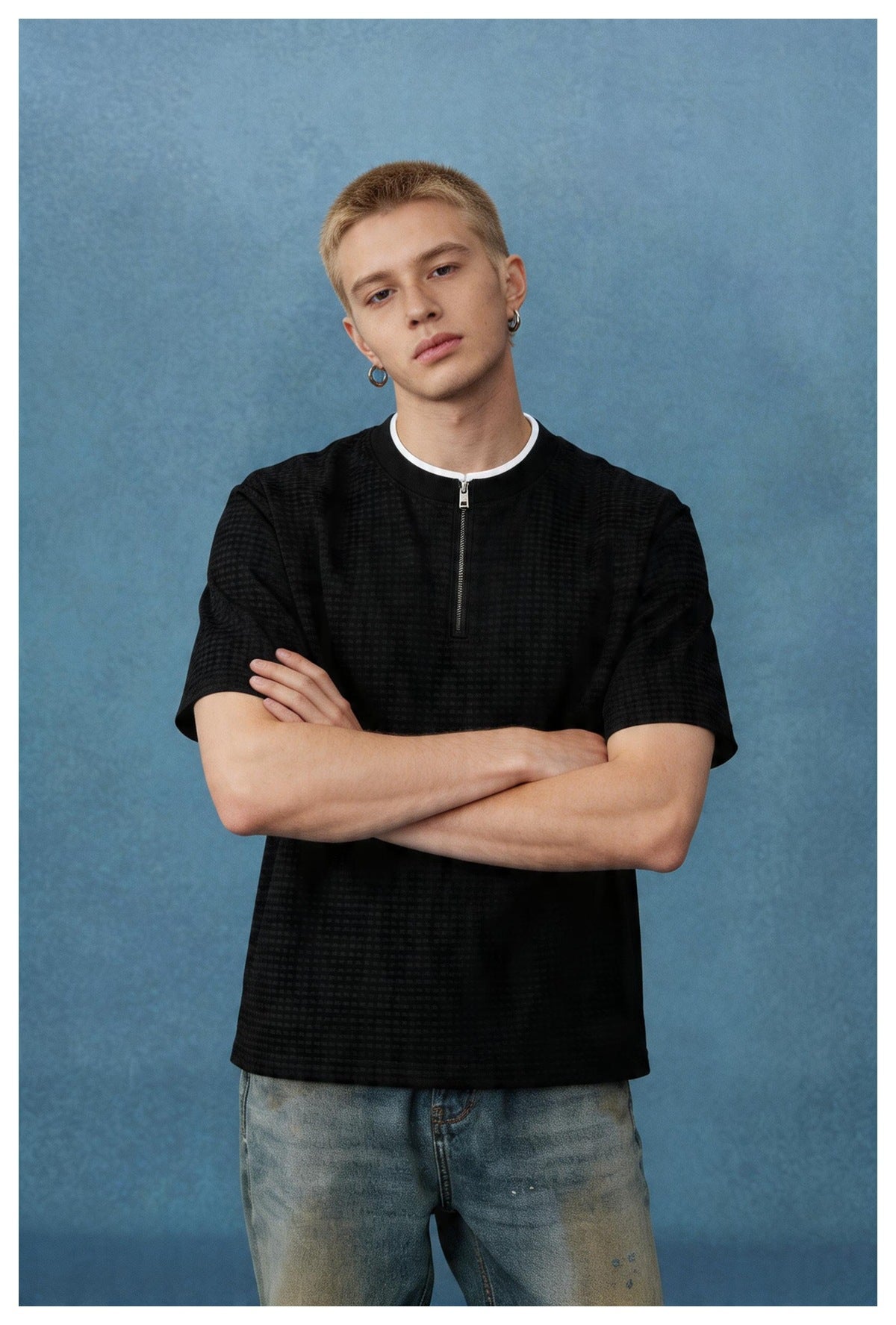 Man wearing a black textured short-sleeve zip-neck T-shirt with white inner collar, arms crossed, with jeans