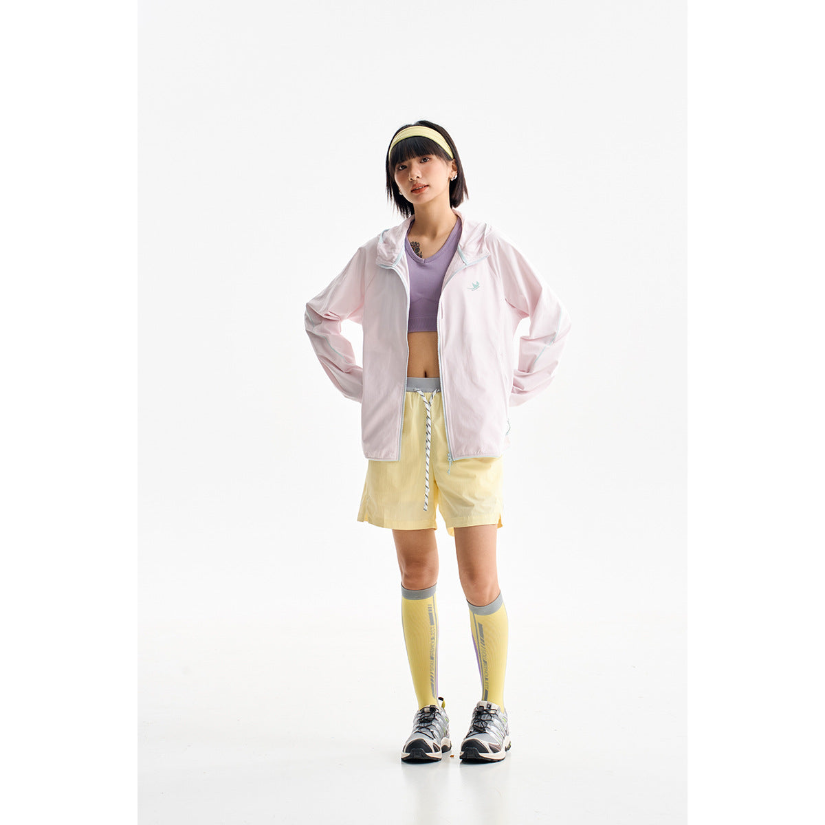 Woman wearing a pale pink hooded zip jacket with aqua trim, a lavender crop top, and yellow shorts.