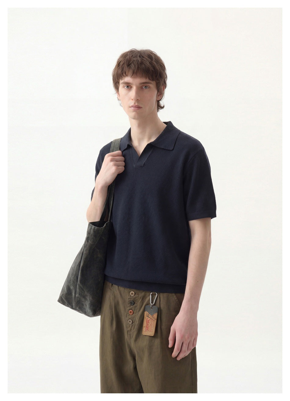 Man wearing a navy knit polo with open V collar, olive trousers and a gray-green tote bag