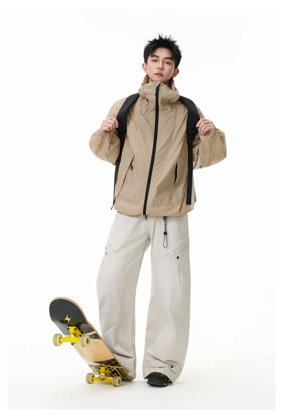 Full-length model wearing a tan high-collar zip jacket and wide light trousers, standing beside a skateboard.