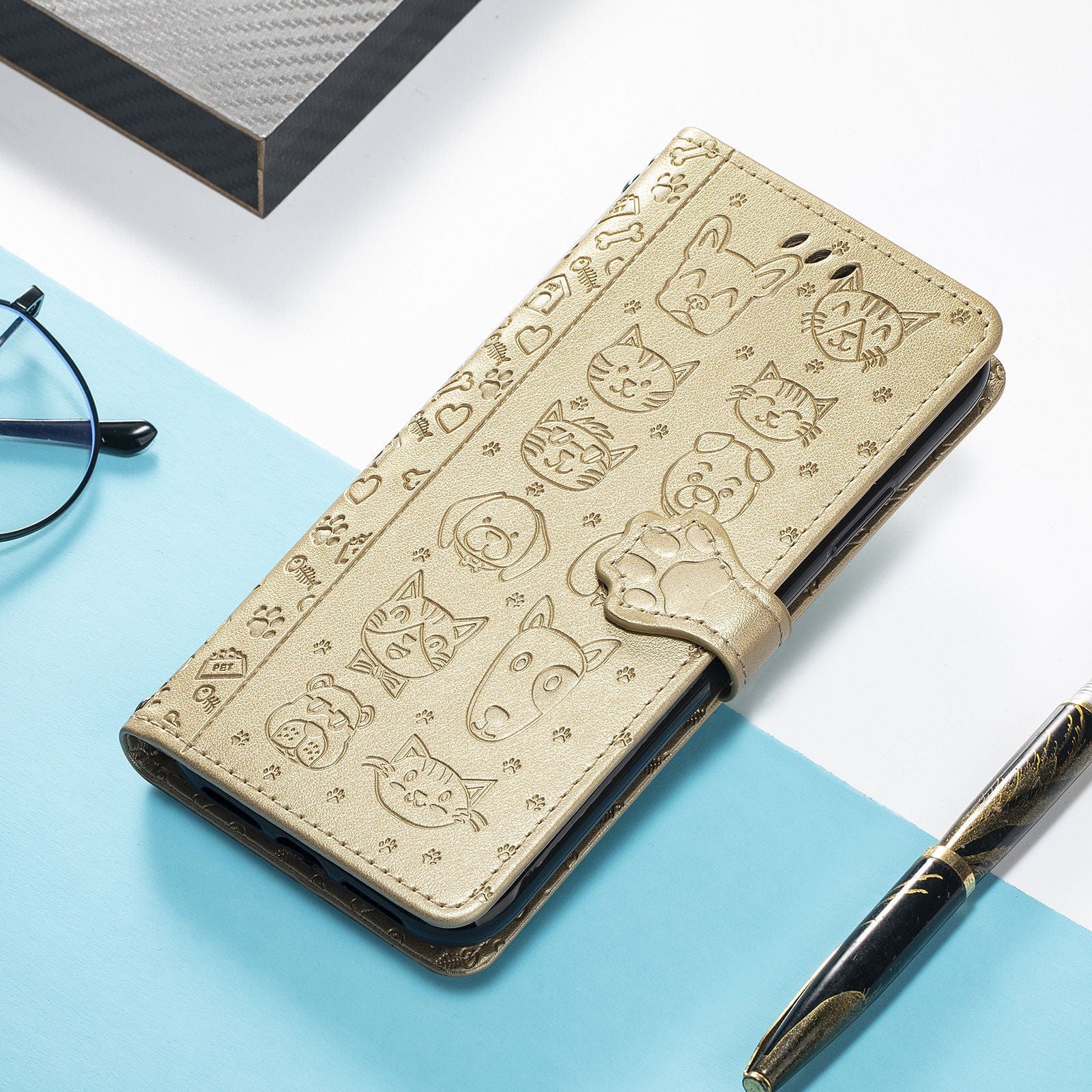 An iPhone 16 Pro Max Wallet Case with a cute cat and dog embossed design in gold lies on a light blue and white surface, accompanied by black-rimmed glasses, a black and gold pen, and a gray book.