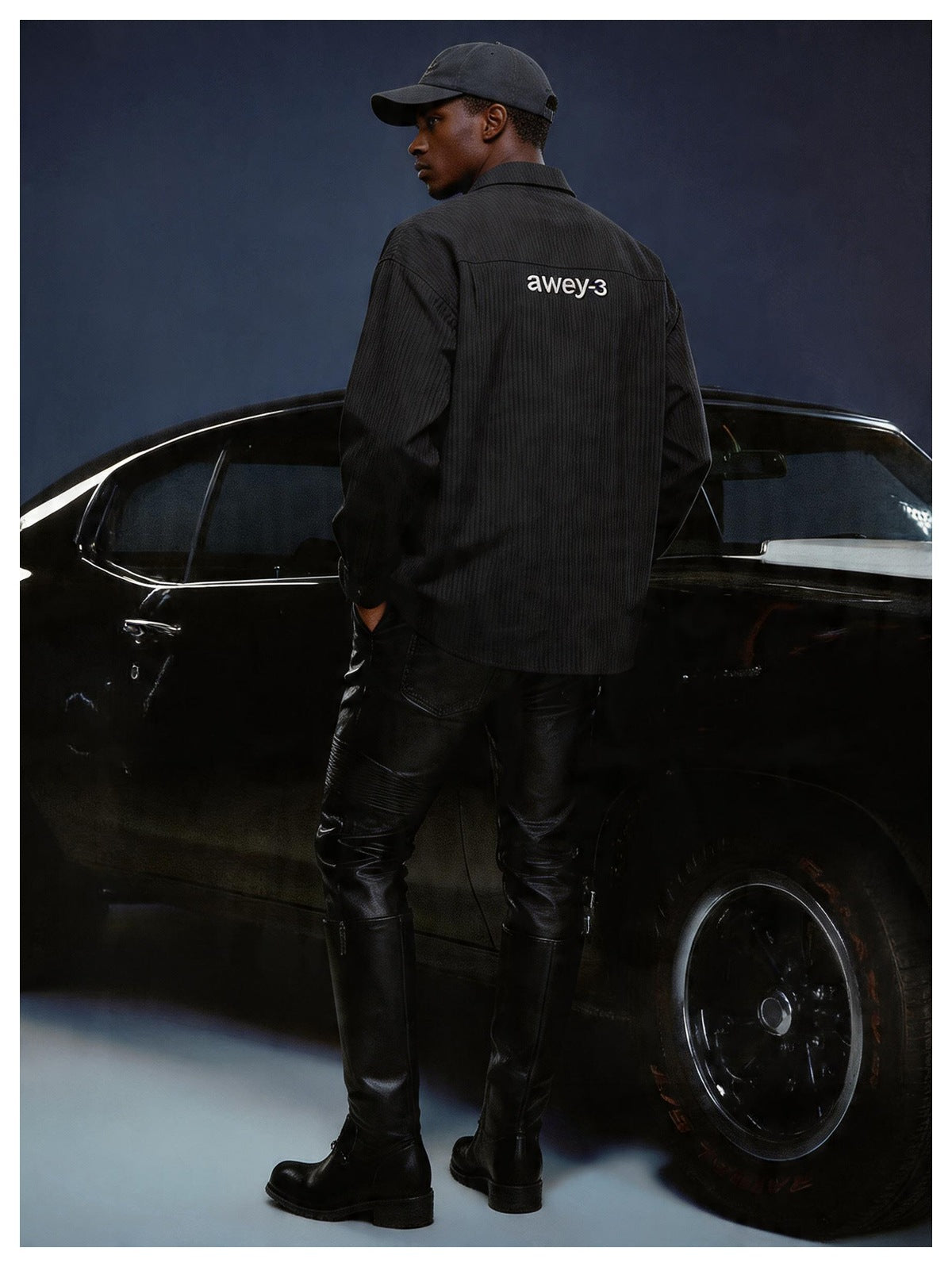 Back view of a man wearing a black striped button-up shirt with embroidered back lettering, a cap, leather pants and boots