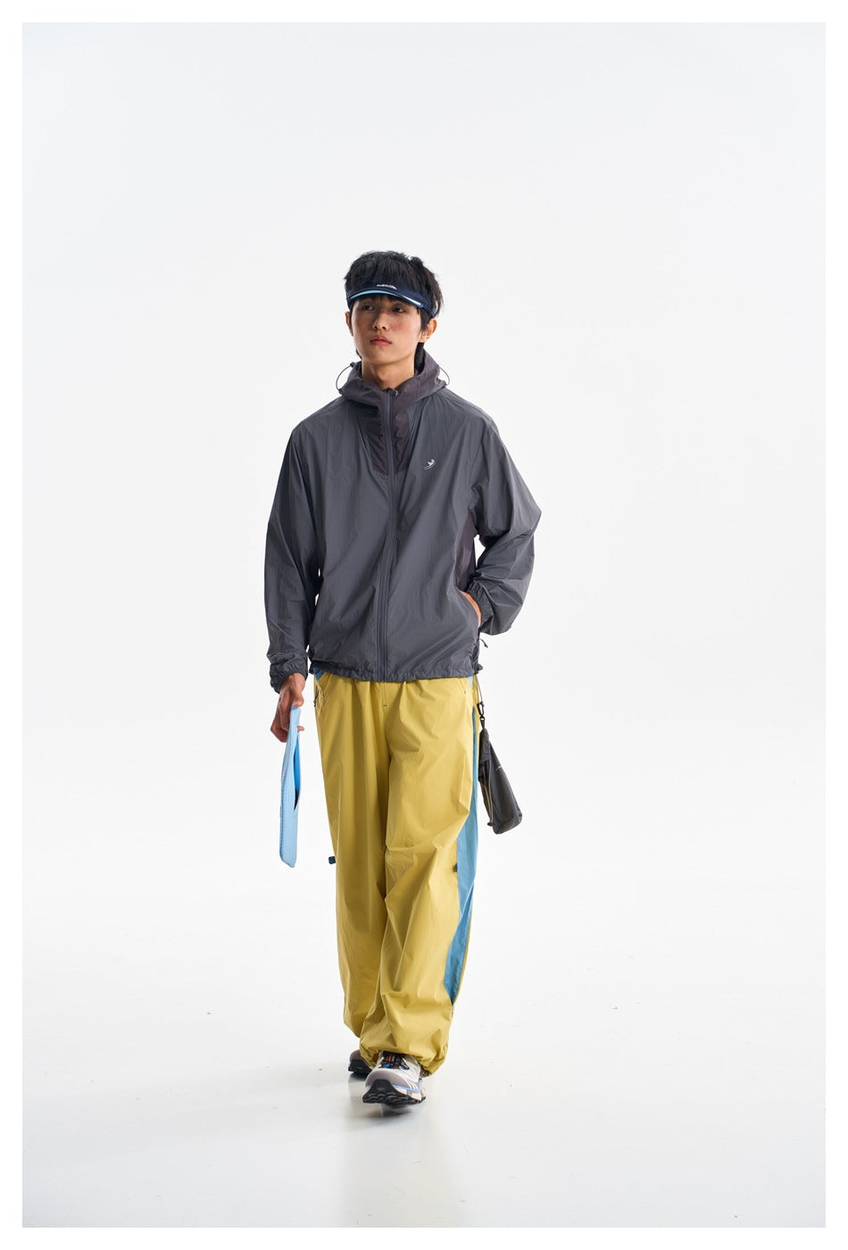 Full-length front view of a person wearing a charcoal hooded jacket and mustard-yellow pants with blue side panels.