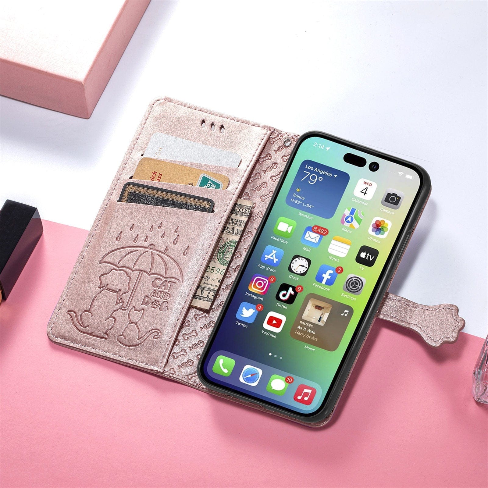A smartphone in a pink PU leather iPhone 16 Pro Max Wallet Case with an embossed cute cat and dog design. The flip cover securely holds bank cards and cash. The phone screen displays various app icons on a pastel pink surface.