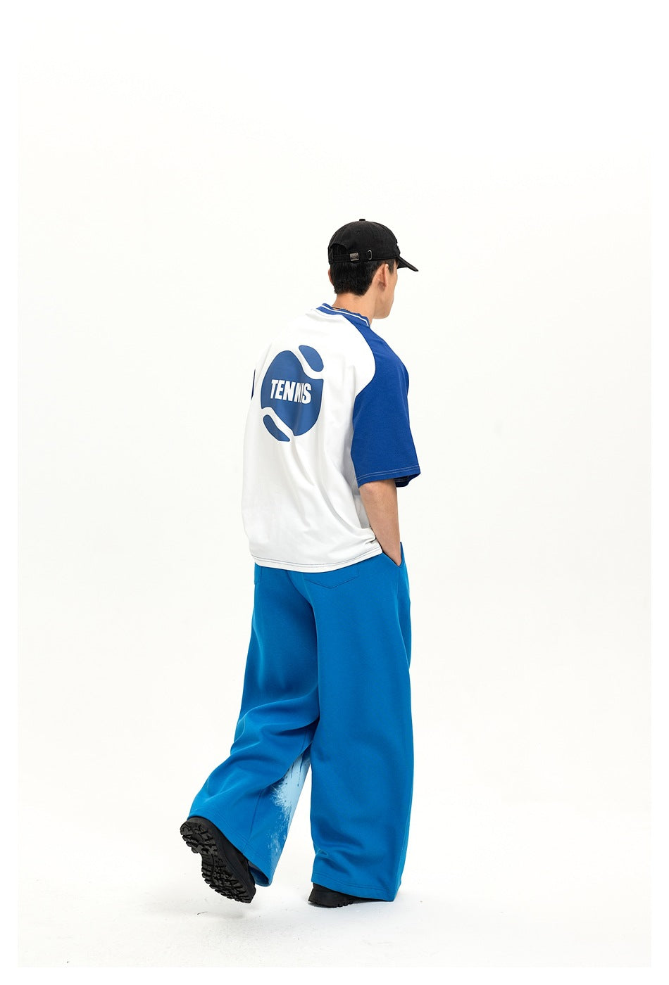 Rear three-quarter view of a model wearing a white-and-blue raglan T-shirt with a large blue tennis graphic and blue pants.