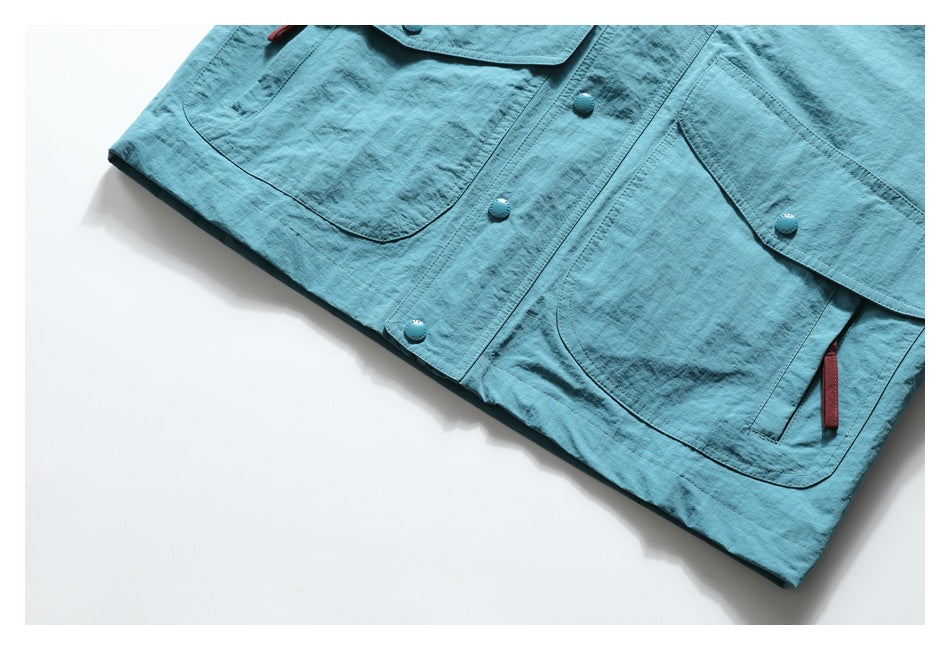 Partial flat-lay view of a teal utility jacket front with large pockets, snap buttons, and red zipper pulls.