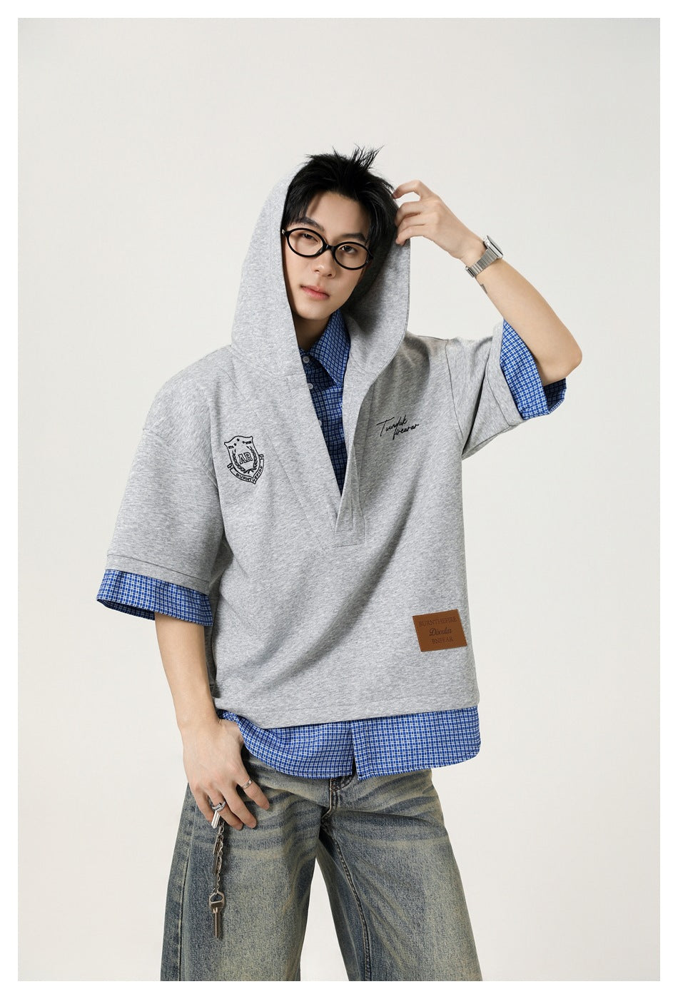 Model wearing a light gray short-sleeve hooded pullover over a blue checked shirt, with wide-leg jeans.