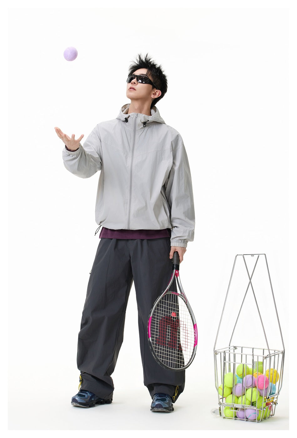 Full-length model wearing a light gray hooded jacket and dark trousers beside a tennis ball basket and racket.