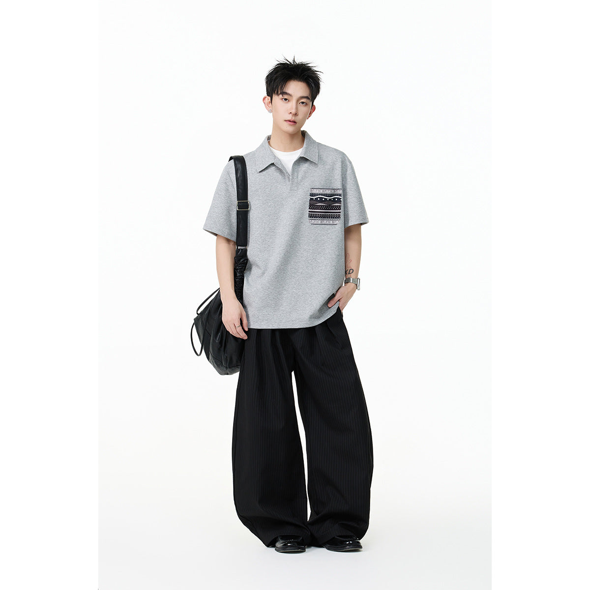 Model wearing a light gray short-sleeve collared polo with a patterned chest pocket and wide black trousers.