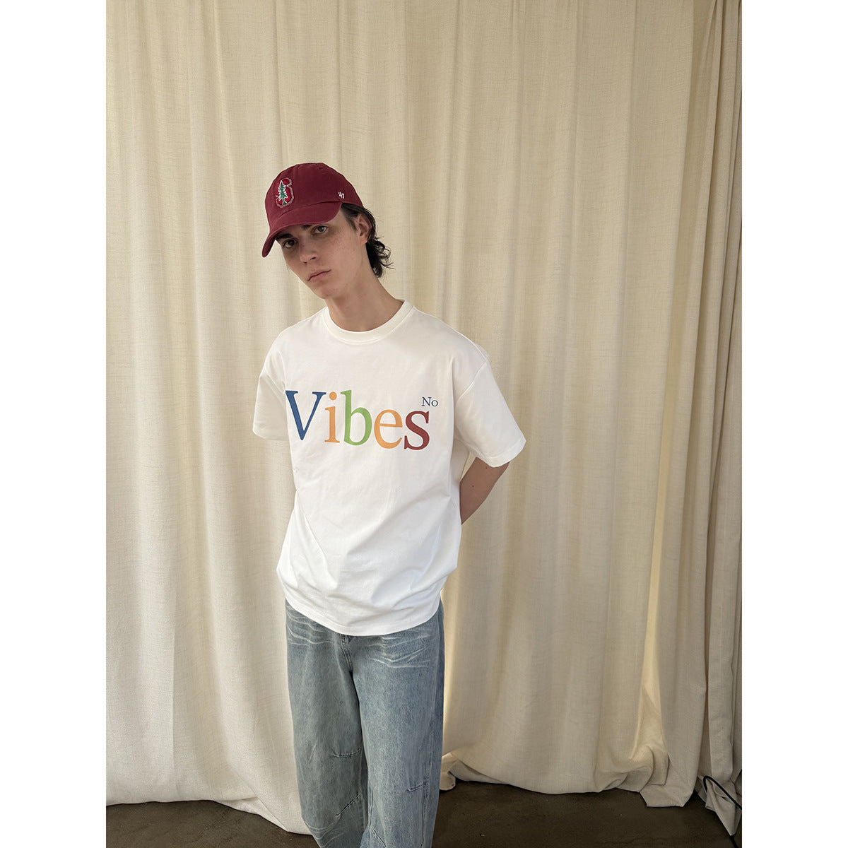 Closer front view of a model wearing a white T-shirt with multicolored “Vibes No” lettering and a burgundy cap.