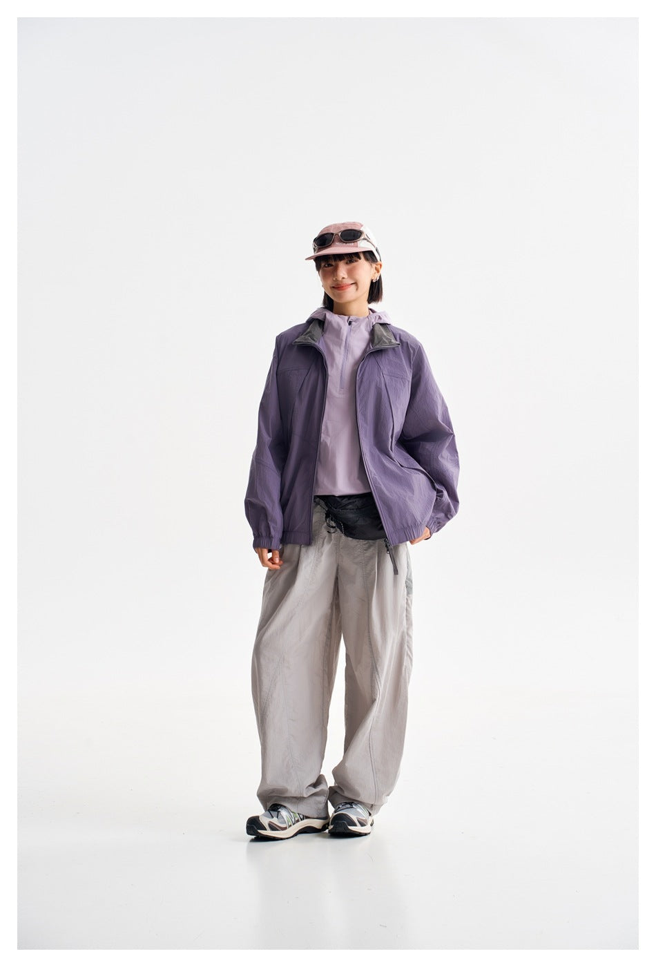 Model wearing a purple zip-front jacket over a light lilac top, with loose gray trousers.