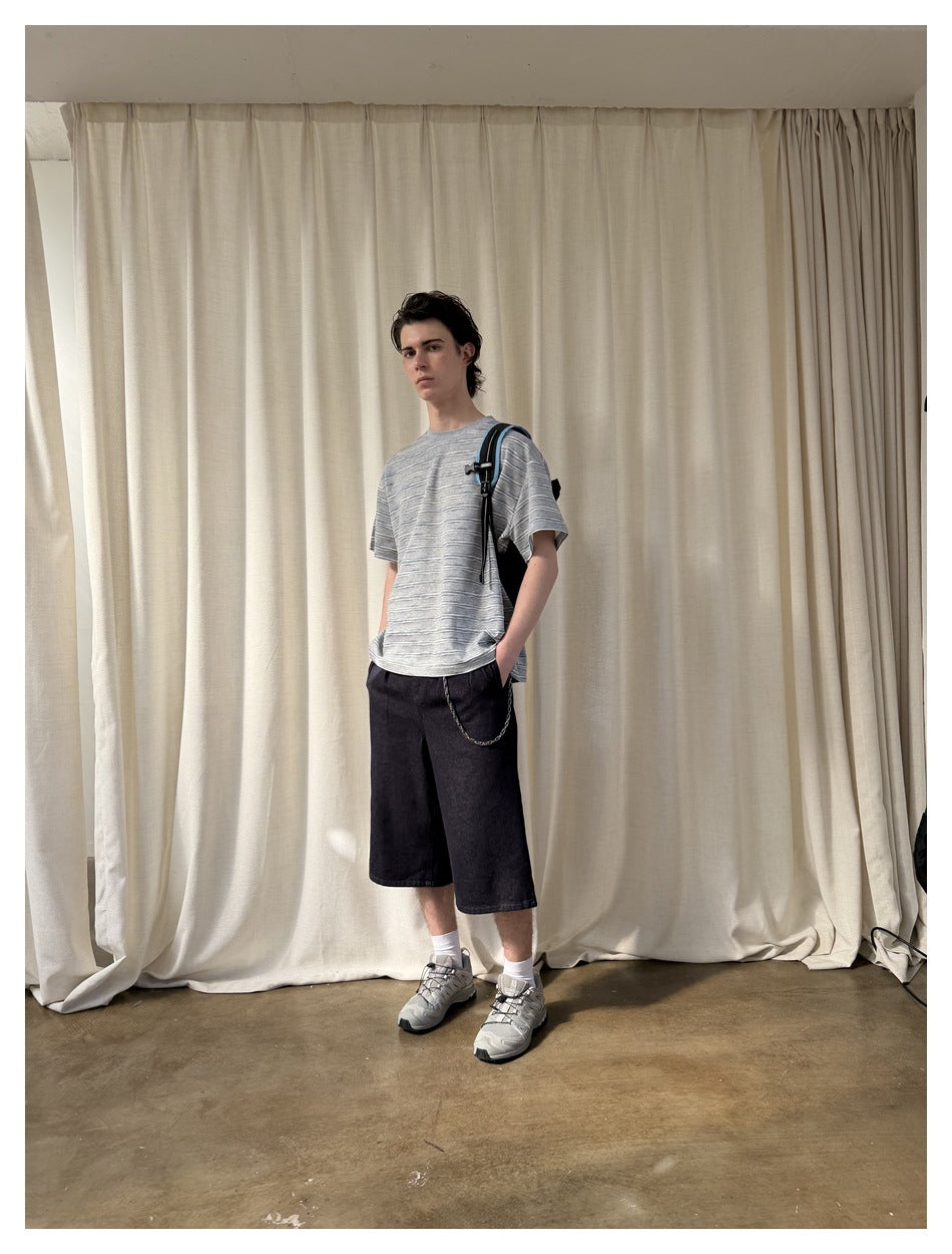 Model wearing a gray striped T-shirt and black knee-length shorts, standing at an angle with a shoulder bag.