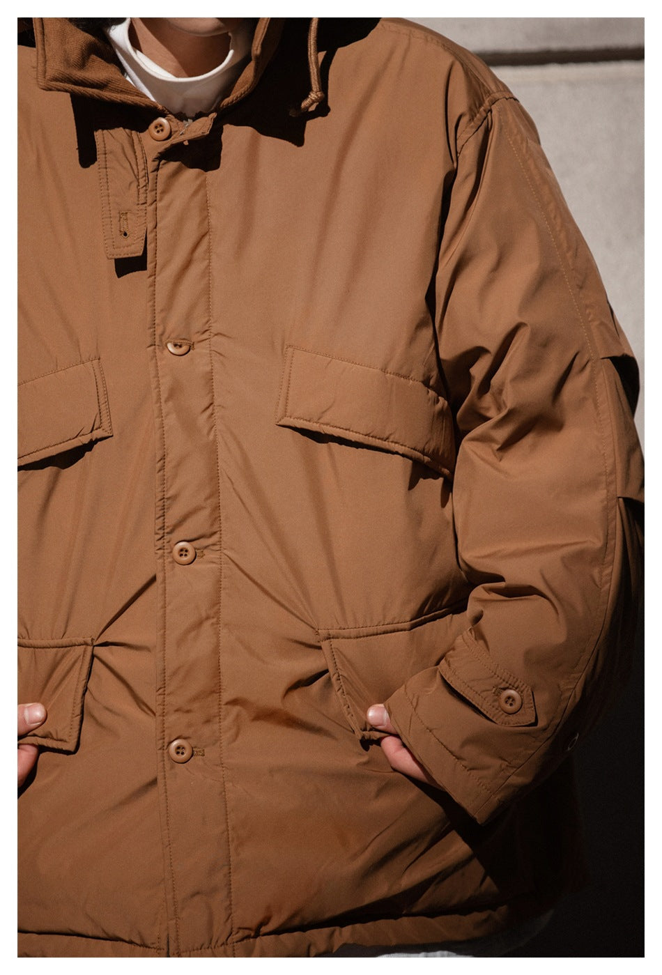 Close-up of a brown jacket showing its button placket, flap pockets, hood drawcord, and cuff tab.
