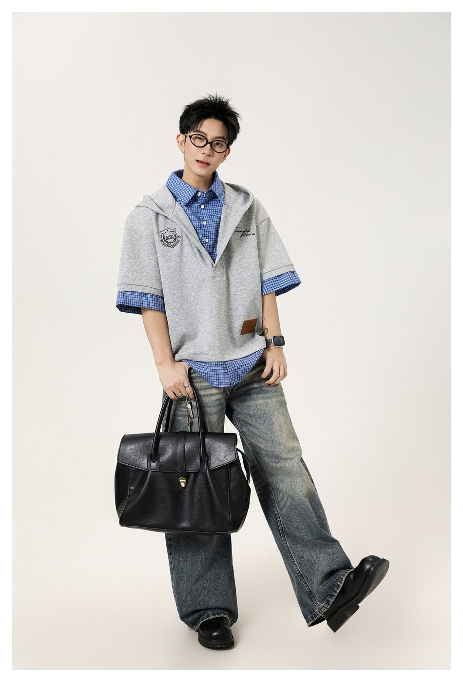 Full-length model wearing a light gray hooded pullover over a blue checked shirt and wide-leg jeans, carrying a black bag.