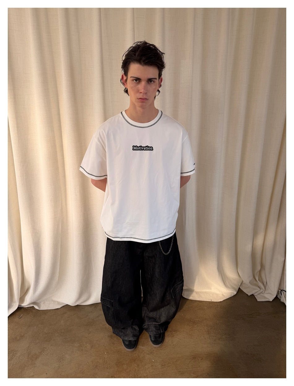 Model wearing a white short-sleeve T-shirt with dark contrast stitching and a small “Motivation” chest print.