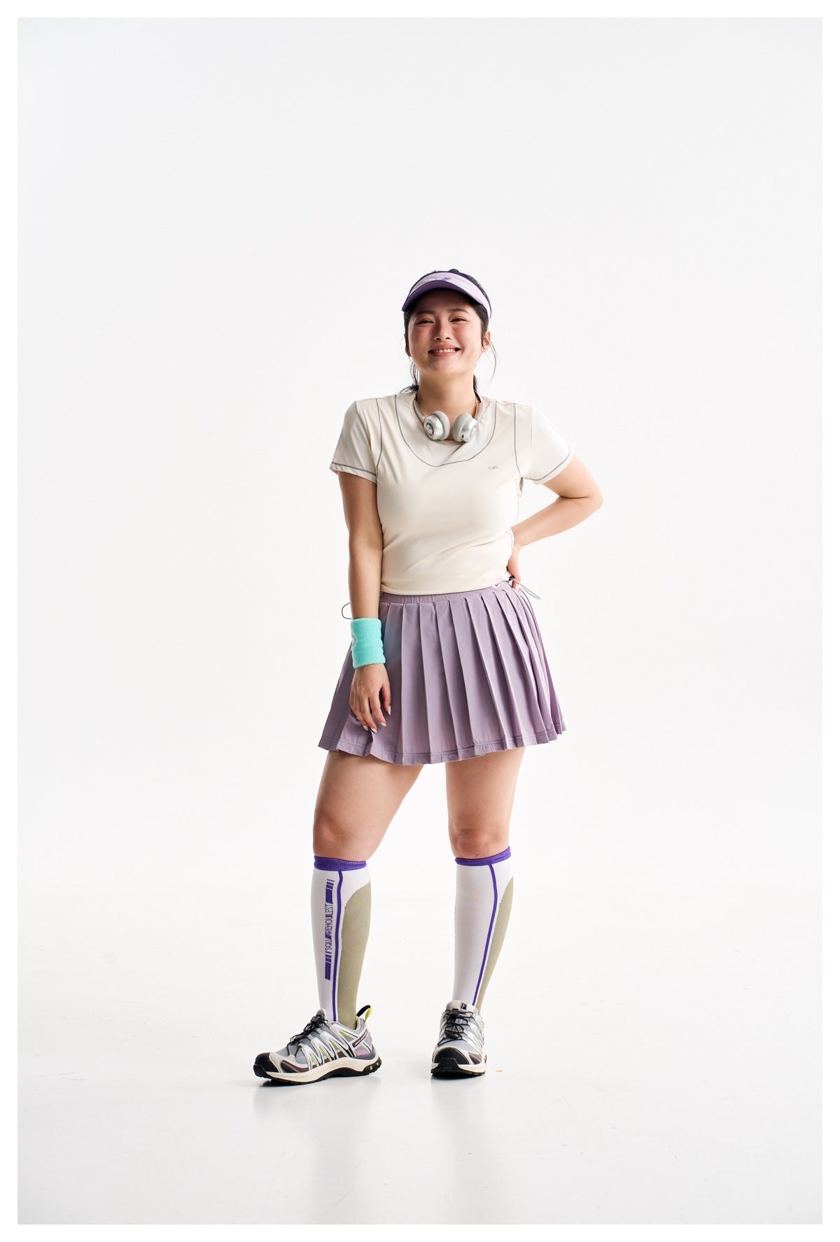 Woman wearing a purple gray pleated mini skirt with a cream top, visor, wristband and tall socks