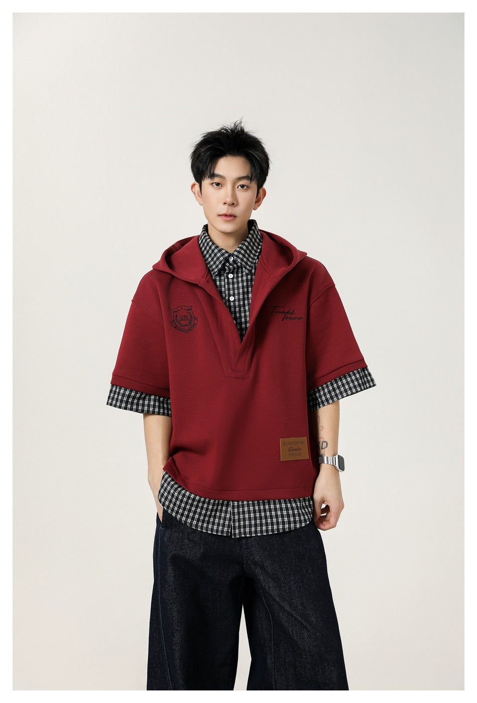 Front view of a model in a burgundy short-sleeve hooded pullover layered over a black-and-white checked shirt.