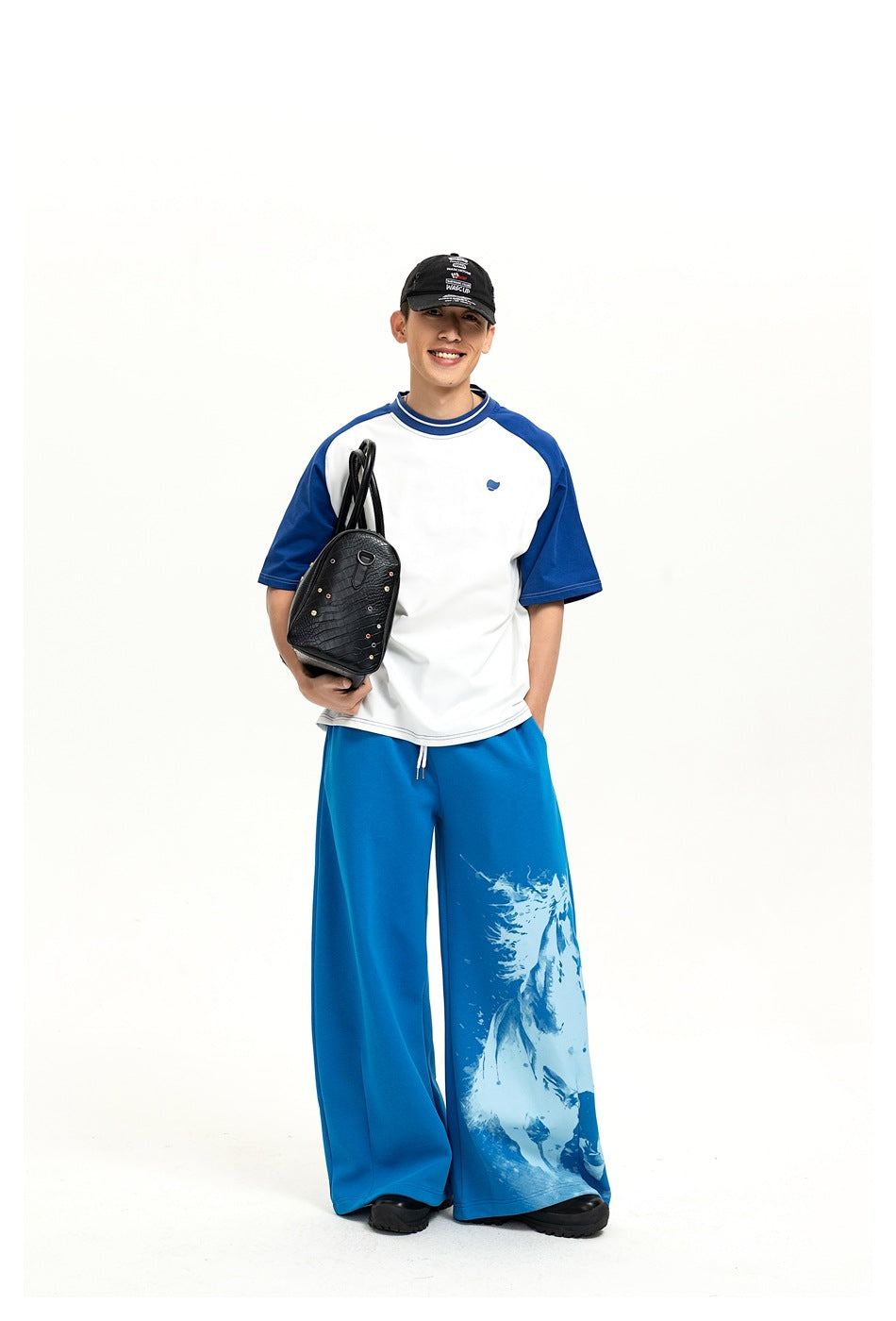 Smiling model wearing a white-and-blue raglan T-shirt and wide-leg blue pants with a pale graphic on one leg.