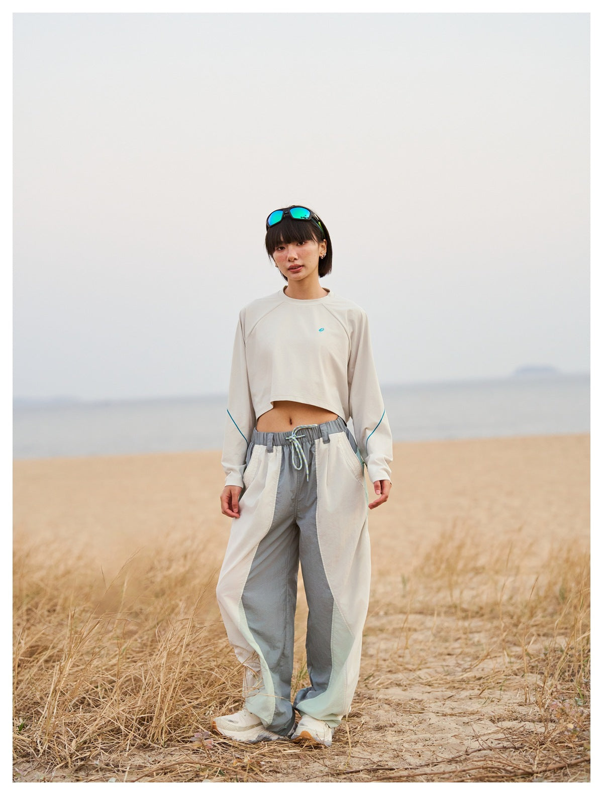 Woman on a sandy beach wearing the white cropped long-sleeve top with teal sleeve piping, styled with wide-leg pants
